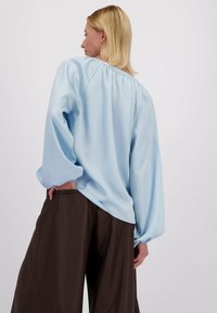 Lichtblauwe blouse met lange mouwen, voorzien van een gathered halslijn en ballonmouwen, gecombineerd met bruine wijde broekspijpen. Vriendelijke stoftextuur.