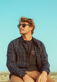 Homme portant des lunettes de soleil noires, une chemise à carreaux bleu marine sur un t-shirt noir, un pantalon beige et une montre numérique, assis à l'extérieur sous un ciel bleu clair.