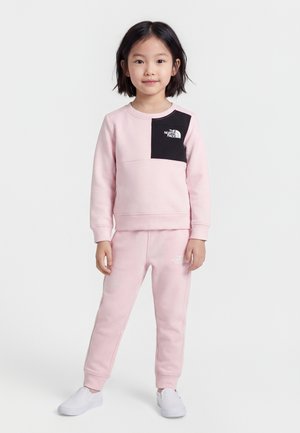 Jeune fille debout portant un sweat-shirt et un pantalon rose clair avec un patch noir et des logos blancs, assortis à des chaussures blanches sans lacets.