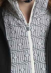 Una giacca con zip caratterizzata da un esterno nero e un rivestimento interno grigio chiaro con un motivo geometrico nero. Tessuto liscio con colletto rialzato.