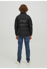 Osoba z kręconymi rudymi włosami, ubrana w czarną kurtkę puchową, niebieskie dżinsy i białe sneakersy, stojąca plecami w stosunku do jednolitego tła.