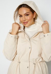 Krémová bunda s kapucí a fleecovou podšívkou, pásek na stažení v pase a patentní knoflíky. Materiál vypadá hladce a má jemnou texturu. Zapínání na zip na přední straně.