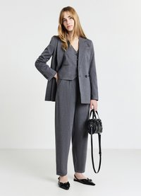 Giovane donna in posa con un completo grigio a righe con gilet e pantaloni, che tiene una borsa nera e indossa ballerine nere a punta su sfondo neutro.