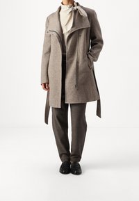 Manteau beige en mélange de laine avec un col montant, une fermeture éclair et des poches latérales, associé à un pantalon foncé et des mocassins noirs.