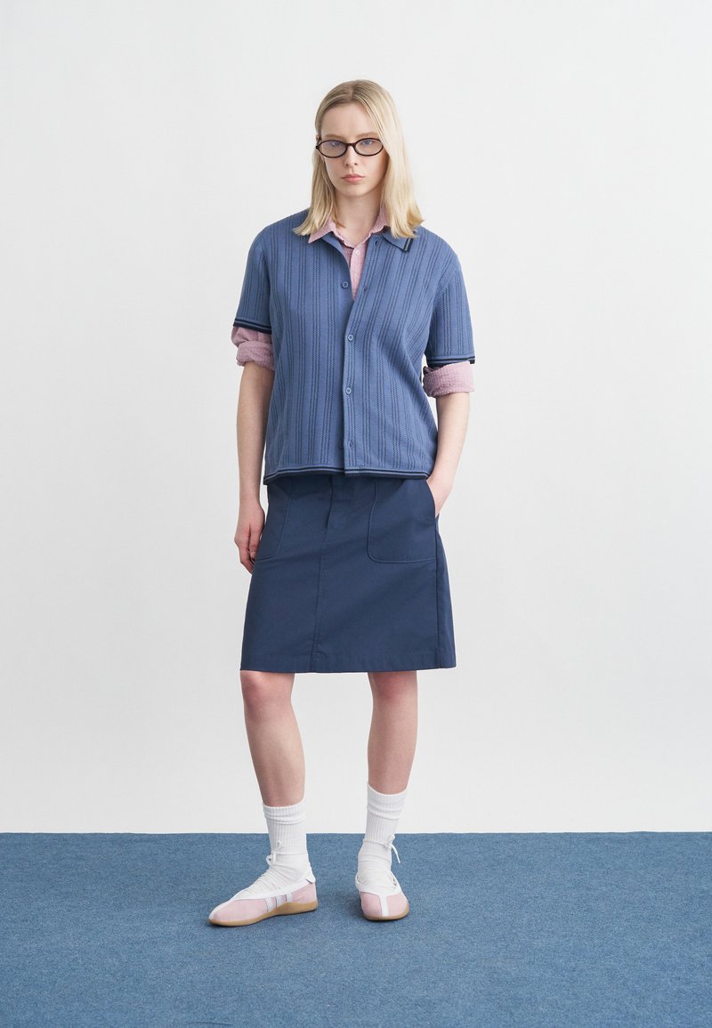 Donna bionda con occhiali, un cardigan blu sopra una maglietta rosa, gonna blu scuro, calzini bianchi e ballerine rosa, in piedi su un tappeto blu.