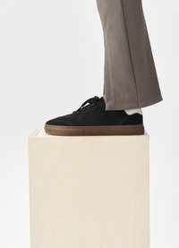 Sneaker din suede negru cu talpă din cauciuc maro, având un design minimalist, închidere cu șireturi și accent alb la toc, așezat pe un bloc din lemn.