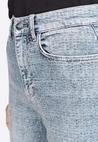 Jeans de mezclilla azul claro con una sutil textura moteada. Presentan un diseño clásico de cinco bolsillos y un cierre de botón plateado.