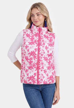 Frau trägt eine weiße Steppweste mit pinkem Blumenmuster, ein weißes Dreiviertelarmshirt, blaue Jeans und blaue Quastenohrringe.