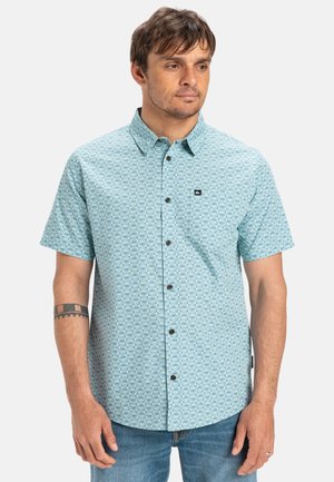 Camisa de botones de manga corta en azul claro con patrón geométrico, que presenta un bolsillo en el pecho y una etiqueta de logo. Textura de tela suave.