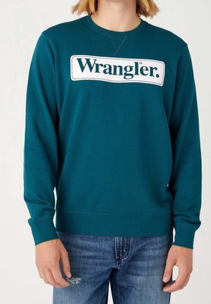 Osoba ubrana w turkusową bluzę Wrangler i niebieskie dżinsy z małą dziurą na lewym udzie, stojąca na jednolitym jasnym tle.