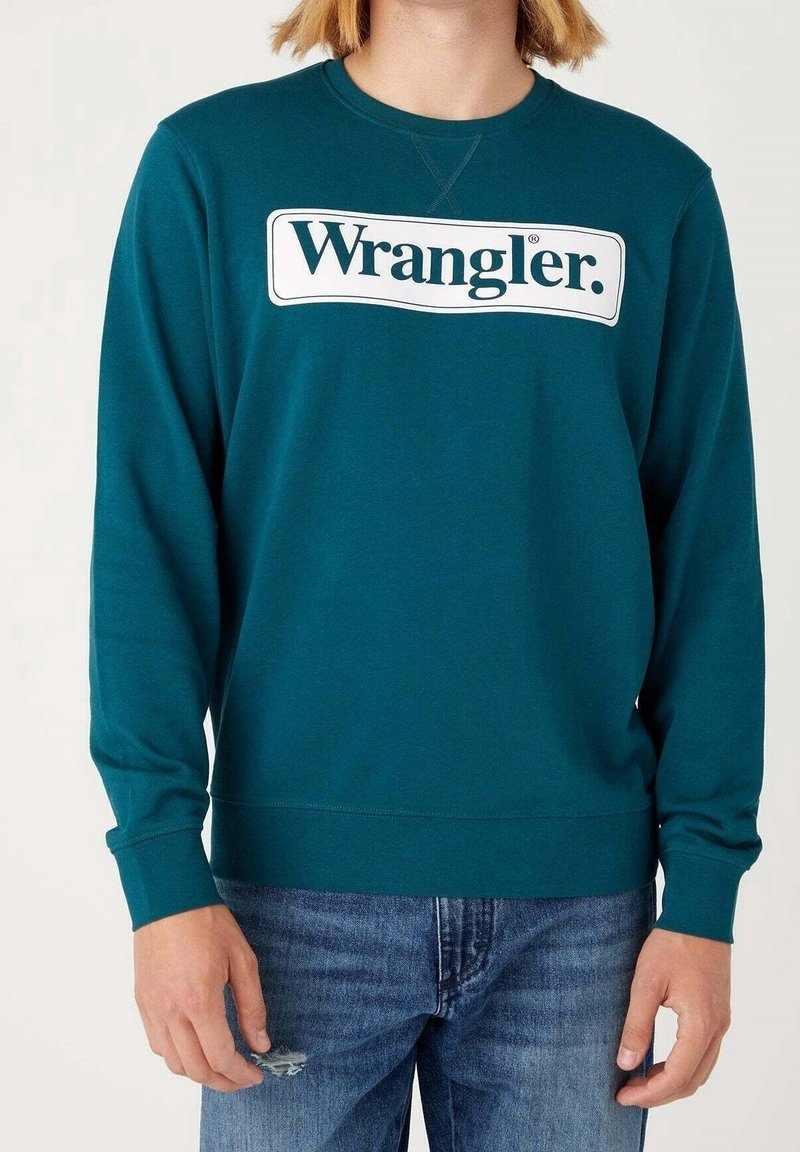 Personne portant un sweat-shirt Wrangler turquoise et un jean bleu avec une petite déchirure sur la cuisse gauche, debout devant un fond clair uni.