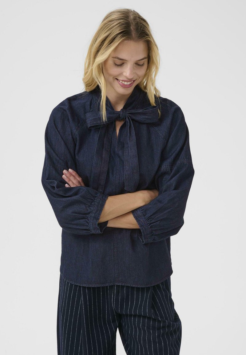 A smiling woman with long blonde hair wears a dark blue blouse tied at the neck and striped pants, standing with arms crossed in a neutral background.