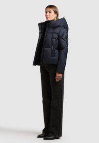 Marineblauwe pufferjack met een hoge kraag, geborduurd ontwerp en grote capuchon. Gekocht met zwarte rechte jeans en zwarte laarzen.