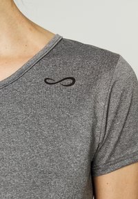 Detailní záběr na osobu v šedém texturovaném tričku s krátkým rukávem, na hrudi blízko výstřihu se nachází černý nekonečný symbol.