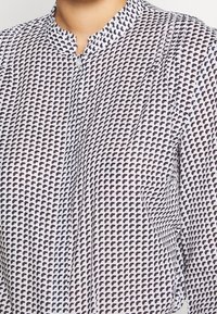 Camisa blanca con un patrón geométrico en azul marino, cuello mao, botonadura y una sutil textura en el tejido. Vista cercana del diseño.