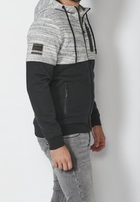 Felpa con zip in tessuto grigio e nero, caratterizzata da una parte superiore testurizzata, due tasche laterali, un cappuccio con cordino e dettagli di branding sulle maniche.