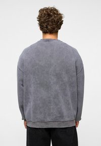 Grijze sweatshirt met een zachte, vervaagde textuur, laagvallende schouders en geribbelde manchetten. De achterkant heeft een schone, simpele uitstraling zonder versieringen.