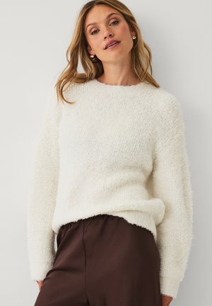 Vrouw in een witte gestructureerde gebreide trui en hoogzitter bruine broek, staand tegen een effen lichtgekleurde muur.