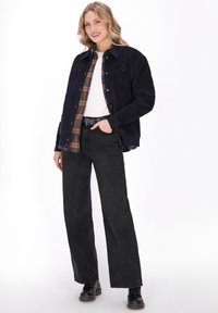 Marine corduroy overhemdjack met knoopsluiting, geruite voering en geborduurde ankerdetail, gecombineerd met hoge zwarte wijde broek.