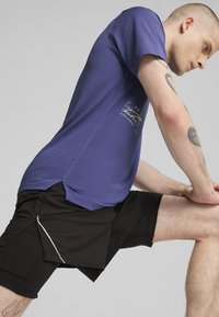 Lilla atletisk t-shirt med en tekstureret overflade, der har et logo. Sammen med sorte shorts, der viser et lagdelt design og reflekterende detaljer.