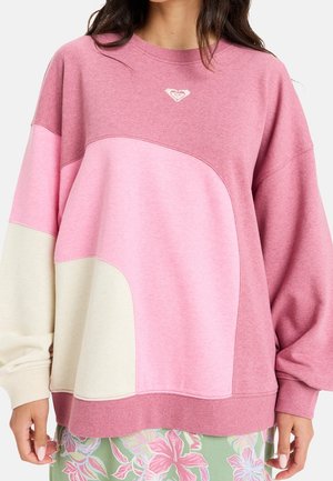 Femme portant un sweat-shirt ample à blocs de couleurs dans des tons roses et beiges avec un petit logo cœur blanc sur la poitrine.