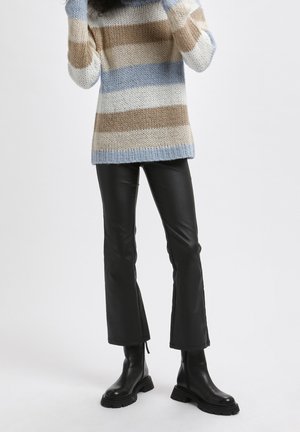 Personne portant un pull en tricot à rayures bleu, blanc et beige, un pantalon en cuir noir évasé et des bottes noires à semelles épaisses, debout sur un fond blanc.