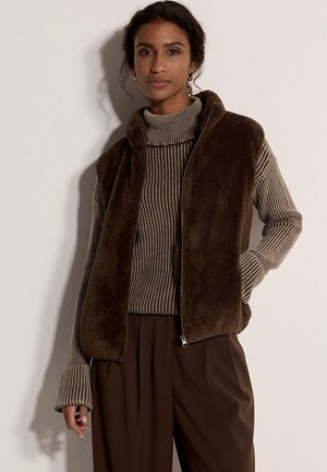 Gilet in pelliccia ecologica marrone con chiusura a zip, indossato sopra un dolcevita a righe marrone. Pantaloni marroni a vestibilità ampia completano l'outfit.