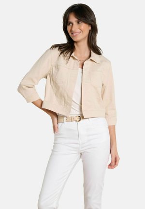 Kobieta uśmiechająca się, ubrana w jasnobeżową krótką kurtkę, biały top, białe spodnie i beżowy pasek, stojąca z jedną ręką na biodrze.