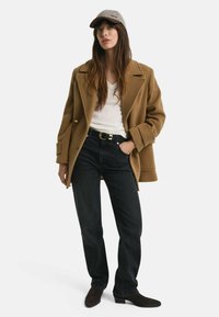 Brauner Wollmischmantel mit goldenen Knöpfen, weißem V-Ausschnitt-Oberteil, dunklen Jeans und grauem Cap. Braune Stiefeletten runden das Outfit ab.