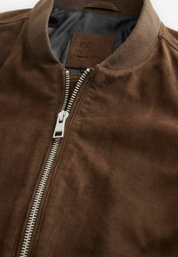 Chaqueta bomber marrón hecha de un material suave y texturizado, con cierre frontal de cremallera y cuello acanalado. Interior forrado con una elegante tela gris.