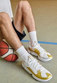 Baskets de sport blanches avec des accents dorés, dessus en maille respirante, semelle texturée, portées avec des chaussettes blanches mi-mollet et un short noir, à côté d'un ballon de basket.