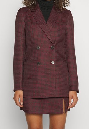 Nainen pukeutunut bordeaux'npunaiseksi ja mustaksi houndstooth-kuvioiseksi kaksiriviseksi bleiseriksi, jonka kanssa hänellä on samasta kankaasta valmistettu etuslissillä varustettu minihame, yhdistettynä mustaan poolopaitaan.