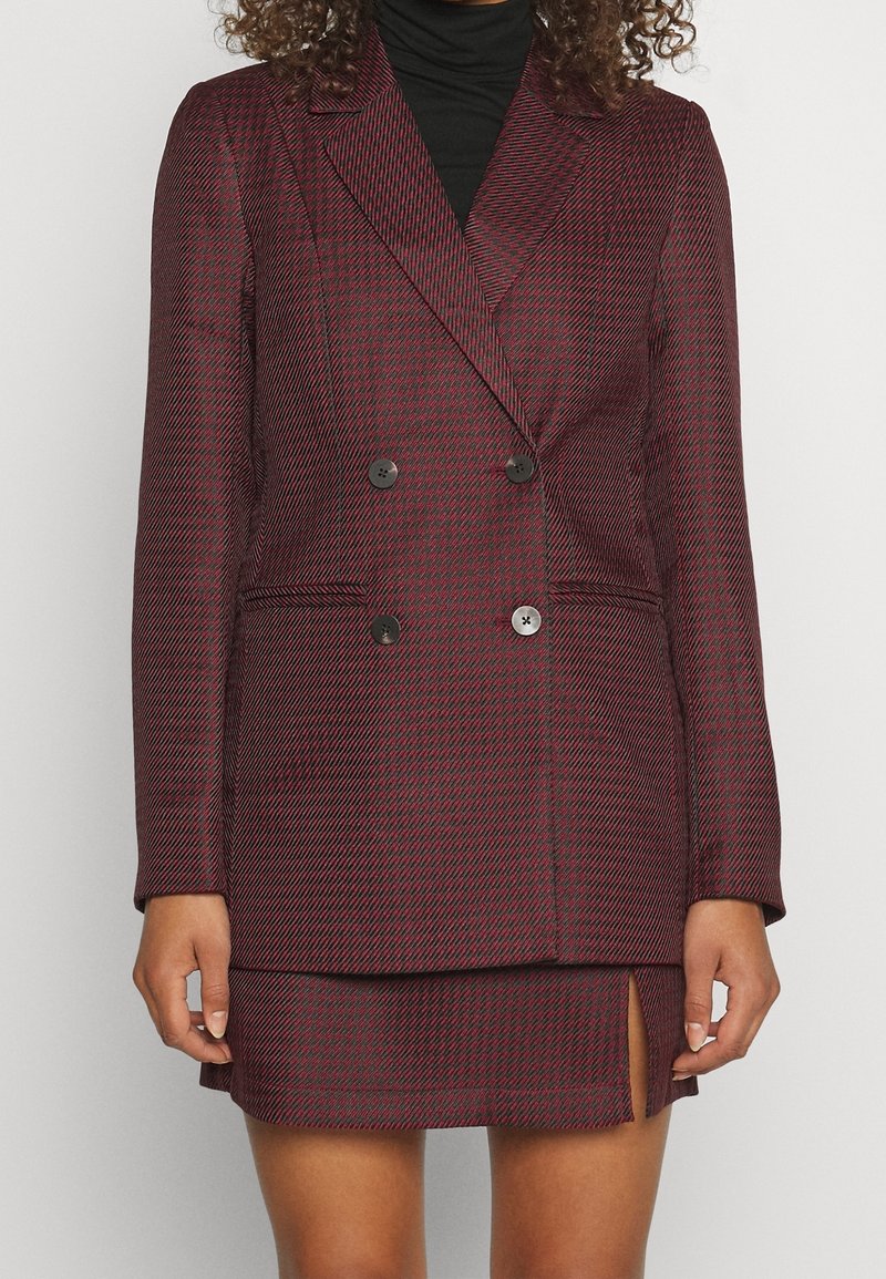 Kvinde iført bourgogne- og sort hundetandsmønstret dobbeltradet blazer med matchende mininederdel med frontslids, kombineret med sort rullekrave.