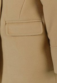 Manteau en mélange de laine beige avec une finition texturée, poche poitrine unique, coupe ajustée et couture détaillée le long des coutures.