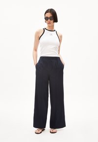 Vit linne med svart kant och logga, i kombination med marinblå vida byxor. Modellen bär svarta runda solglasögon och platta sandaler. En enkel bakgrund.