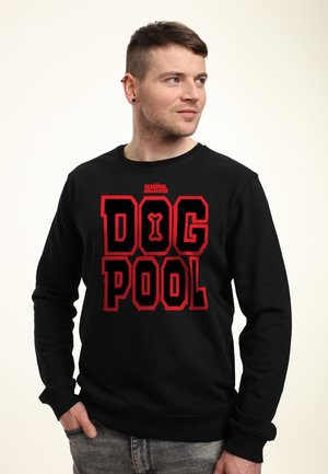 Młody mężczyzna z krótkimi włosami, ubrany w czarną bluzę z czerwonym napisem "Dog Pool" i małym napisem "Deadpool Wolverine" powyżej, patrzący na bok.