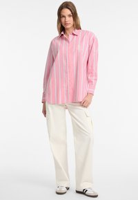 Camicia a righe rosa con linee verticali blu, maniche lunghe e vestibilità rilassata, abbinata a pantaloni cargo crema e scarpe casual.