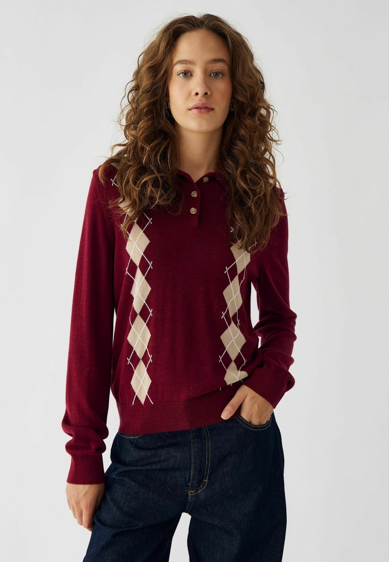 Polo de manga larga burdeos con tres botones y patrón argyle cremoso. Tejido suave y texturizado con puños acanalados. Combinado con jeans oscuros.