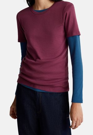 Person trägt ein bordeauxfarbenes Kurzarmhemd über einem blauen Langarmhemd und dunkle Hosen, die vor einem schlichten Hintergrund steht.