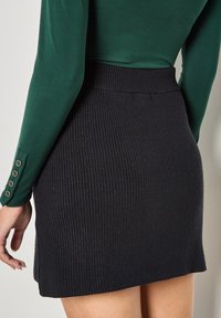 Gonna mini nera a costine con vita alta. Presenta una fessura laterale ed è abbinata a una maglietta a maniche lunghe verde aderente con dettagli in bottone.