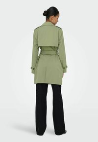 Femme portant un trench-coat ceinturé vert olive et un pantalon large noir, debout de dos, coiffée d'un chignon et portant des boucles d'oreilles créoles.