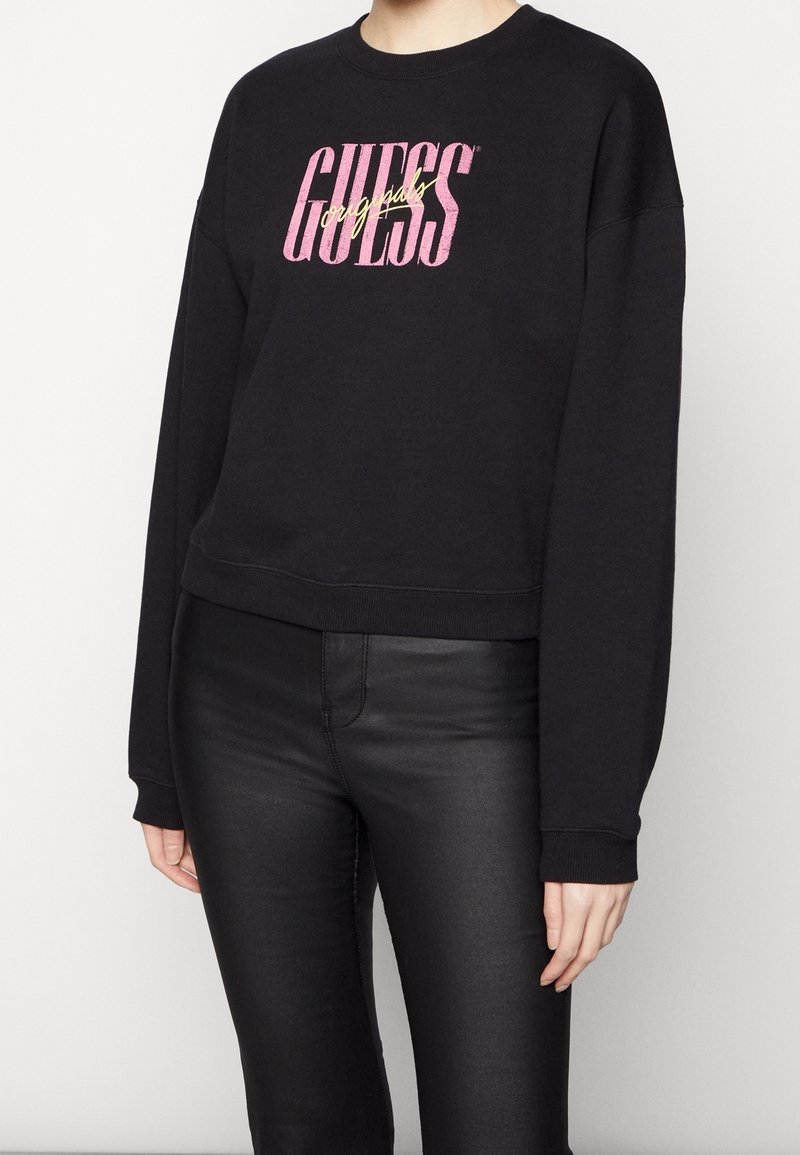 Svart kort sweatshirt med långa ärmar, med en djärv rosa och guld "GUESS" grafisk text över framsidan. Slät bomullstyg.