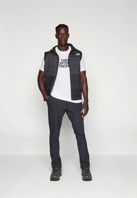 Mann iført sort polstret vest, hvid t-skjorte fra The North Face, sorte bukser og mørke vandresko, stående med hendene i lommene.