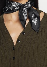 Olivgrön ribbad tröja med djup V-ringning och tre svarta knappar; accentuerad med en svart silkscarf med vita mönster knuten runt halsen.