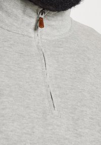 Närbild av en ljusgrå stickad zip-sweater med ett dragkedjehandtag i brunt läder, buren av en person med skägg.