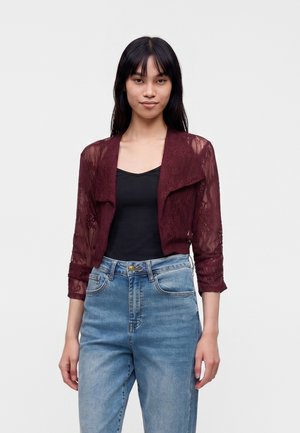Jeune femme portant un cardigan en dentelle bordeaux transparent sur un haut noir et un jean bleu taille haute, debout devant un fond blanc uni.