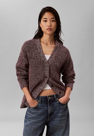 Jeune femme aux longs cheveux foncés porte un cardigan en maille bordeaux par-dessus un crop top blanc et un jean bleu, debout les mains dans les poches.