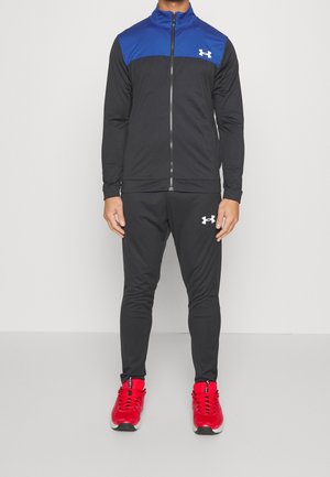 Homme portant une veste de sport zippée noire et bleue, un pantalon de sport noir avec un logo blanc et des chaussures de sport rouge vif, debout contre un mur blanc.