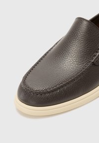 Gros plan sur un mocassin en cuir marron foncé à surface texturée et semelle en caoutchouc crème sur un fond clair uni.
