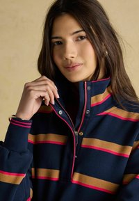 Pulover marin cu dungi orizontale în aurie și roz. Prezintă un guler cu fermoar și capse și manșete cu textură ribbit. Textură moale a materialului.